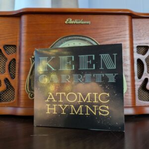 a compact disc in a paper wallet printed with the words KEEN GARRITY ATOMIC HYMNS stands against an old-fashioned record player