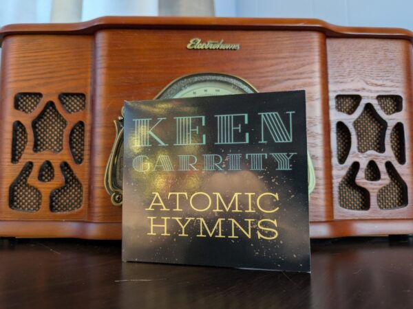 a compact disc in a paper wallet printed with the words KEEN GARRITY ATOMIC HYMNS stands against an old-fashioned record player