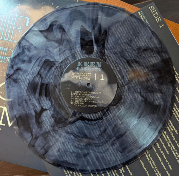 The translucent vinyl disc of Atomic Hymns, which features a black marble splatter pattern. The disc sits on top of the printed lyric sleeve and outer sleeve.