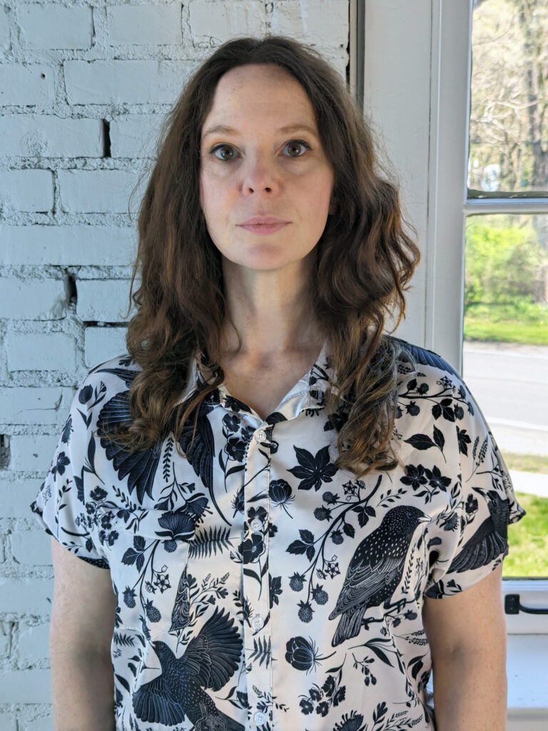 a woman stands against a white brick wall and window. She is wearing a white collared blouse emblazoned with a black starling pattern. Her auburn hair is worn down with loose curls.