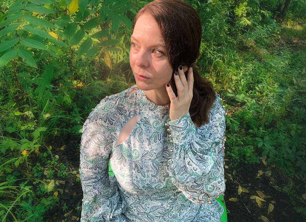 Keen Garrity, wearing a paisley dress, touching hair, seated among leaves in the woods
