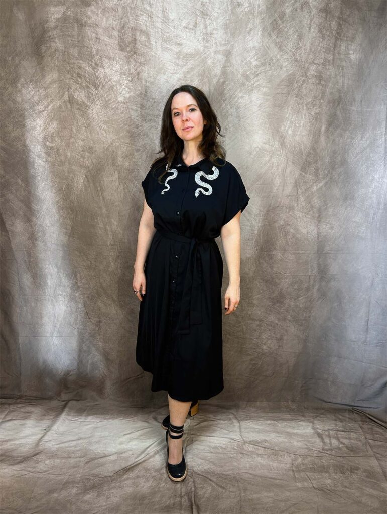 Keen Garrity, standing in black dress with white snake embellishment on shoulders, in front of a brown backdrop with beams on light on either side