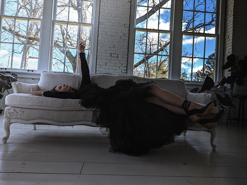 Keen Garrity, lying on a white sofa in front of tall windows, reaching with one arm toward the ceiling